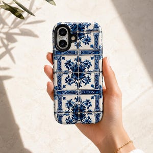 May include: A white and blue floral patterned phone case. The case features a repeating design of blue flowers and geometric shapes on a white background. The phone case is held in a hand.