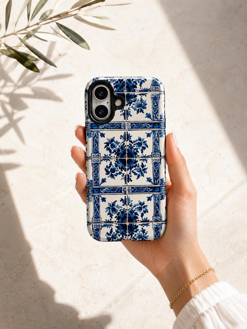 May include: A white and blue floral patterned phone case. The case features a repeating design of blue flowers and geometric shapes on a white background. The phone case is held in a hand.