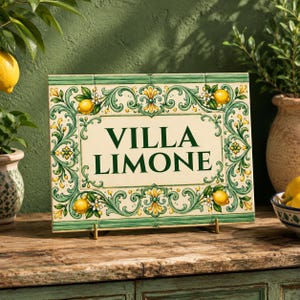 May include: A rectangular ceramic sign with a cream-coloured centre featuring the dark green text VILLA LIMONE. The border is decorated with green scrollwork, yellow lemons, and small white flowers on a light green background.