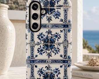 May include: A white and blue floral patterned phone case. The case features a tile design with blue floral motifs and decorative borders. The phone case has a camera array on the back. The background includes a coastal scene and a book titled "Coastal Living."