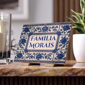 Può includere: Una piastrella decorativa con le parole "FAMILIA MORAIS" in blu. La piastrella presenta un motivo floreale blu su sfondo crema ed è esposta su un supporto in legno. È un elemento decorativo per la casa.