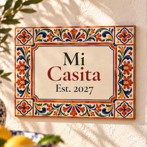 Könnte beinhalten: Ein rechteckiges Wand-Schild aus Keramik mit cremefarbener Mitte, das den dunkelgrünen Text Mi Casita sowie Est. 2027 in Rot und Grün zeigt. Der Rand ist mit einem bunten Blumenmuster in Orange, Blau, Grün und Gelb verziert.