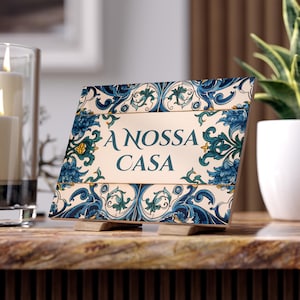 Puede incluir: Una baldosa decorativa con las palabras "A NOSSA CASA" en verde azulado, enmarcada por un diseño floral azul y blanco. La baldosa se apoya en dos pequeños soportes de madera, adecuada para exhibir en una mesa o estante.