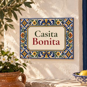 Puede incluir: Una placa de pared rectangular de cerámica con un centro de color crema que presenta el texto Casita Bonita en letras de color verde oscuro y rojo. El borde está decorado con un vibrante patrón floral en tonos azul, amarillo, naranja y verde.
