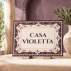 以下が含まれることがあります： クリーム色の中央に濃い紫色の文字で「CASA VIOLETTA」と書かれた長方形のセラミック製看板。縁取りには複雑な金と紫のスクロール模様、小さなピンクの花のアクセント、そして濃い紫色の外枠が施されています。