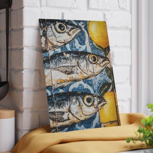 May include: A rectangular wooden art piece featuring three fish illustrations with large eyes, set against a blue and white patterned background. Yellow lemons are also depicted. The artwork is leaning against a white brick wall.