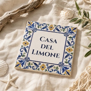 Könnte beinhalten: Quadratische Keramikfliese mit den Worten "CASA DEL LIMONE" in Marineblau. Die Fliese hat einen weißen Hintergrund mit einem dekorativen Rand aus blauen und gelben Blumen- und Blättermustern. Muscheln und Treibholz im Hintergrund.