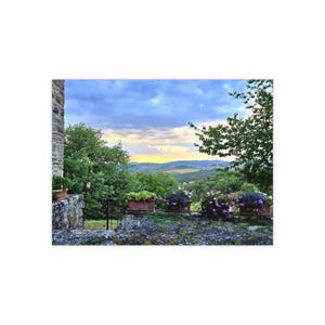 May include: A stone terrace features several rectangular terracotta planters filled with vibrant purple and pink flowers. The terrace overlooks a lush green landscape of rolling hills under a cloudy sky with a soft golden sunset glow on the horizon.