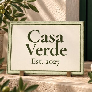 May include: A rectangular sign with a cream-coloured background and a thin green border. The text "Casa Verde" is printed in large, dark green serif letters, with "Est. 2027" in smaller dark green letters centred underneath the main text.
