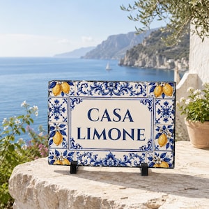 May include: A rectangular slate sign featuring the text CASA LIMONE in dark blue lettering. The border is decorated with a white and blue floral pattern and yellow lemon illustrations on a white background, supported by two black metal stands.