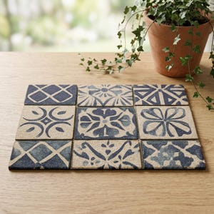 May include: Nine square coasters with a blue and beige tile design. Each coaster features a different geometric or floral pattern. The coasters are arranged on a wooden surface, with a potted plant in the background.