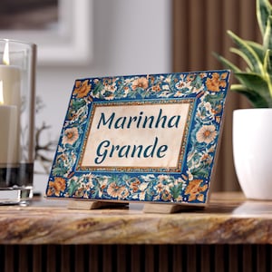 May include: A decorative tile with the words "Marinha Grande" in a vintage font. The tile features a blue border with floral and leaf patterns in orange and cream colors. It rests on two small wooden supports.