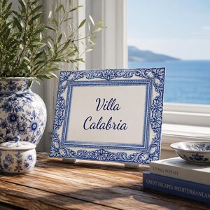 May include: A ceramic sign with the words "Villa Calabria" in navy blue script. The sign has a white background and a detailed blue floral border. It is displayed on a wooden surface with other ceramic items and books.