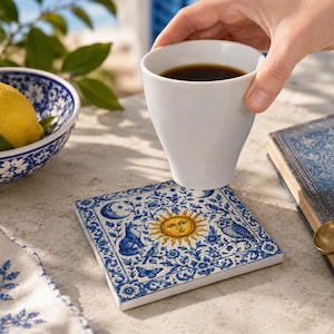 May include: A square ceramic coaster featuring a central yellow sun with a face, surrounded by blue and white floral patterns, a fox, an owl, and a crescent moon. The coaster is positioned beneath a white ceramic cup filled with dark coffee.