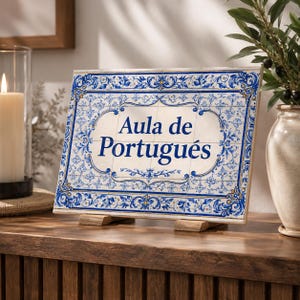 Può includere: Un cartello rettangolare in piastrelle con le parole "Aula de Português" in blu. Il cartello ha uno sfondo bianco con motivi floreali blu ed è esposto su un supporto di legno. Il cartello è su una superficie di legno.
