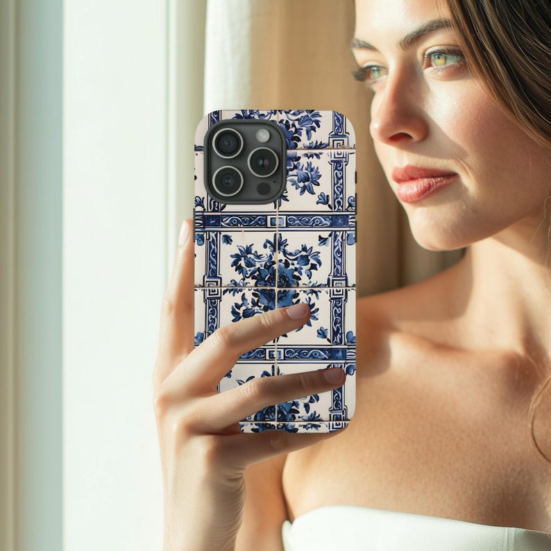 May include: A smartphone case featuring a blue and white floral tile design. The case has a square pattern with a repeating floral motif. The case is held in front of a window, showcasing the intricate details and color contrast.