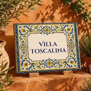 May include: A rectangular ceramic sign featuring the text VILLA TOSCALINA in dark blue lettering. The sign is decorated with a colourful border of yellow flowers, green leaves, and blue scrollwork patterns on a white background.