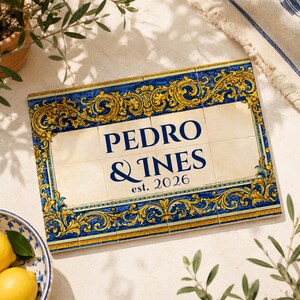 Könnte beinhalten: Eine rechteckige Wandplatte aus zwölf Keramikfliesen mit einem cremefarbenen Zentrum, das den dunkelblauen Text "PEDRO & INES est. 2026" trägt, umgeben von einem dekorativen Rand aus komplexen goldenen und blauen Rankenmustern.
