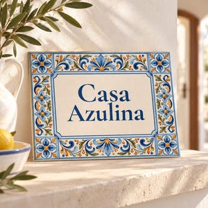Puede incluir: Una placa de pared rectangular de cerámica con el texto Casa Azulina en letras azul oscuro. La placa está decorada con un borde pintado a mano de flores azules, hojas verdes y detalles en naranja sobre un fondo de color crema.