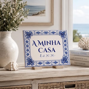 May include: A rectangular ceramic plaque with a blue and white floral border and the text "A MINHA CASA - Est 2026". The plaque is displayed on a wooden stand, with a vase of flowers and books in the background.