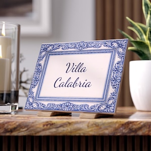 May include: A rectangular sign with the words "Villa Calabria" in elegant script. The sign has a white background with a blue floral border and is propped up on two wooden supports. A candle and plant are in the background.
