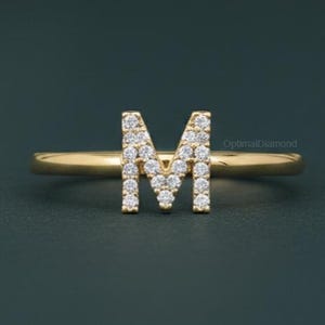May include: A gold ring featuring the letter "M" encrusted with small, sparkling diamonds. The ring has a slender band and the letter is prominently displayed in the center. The background is a dark teal color.