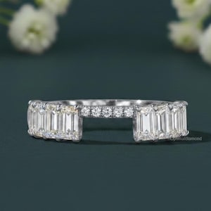 May include: A silver ring featuring emerald-cut diamonds and smaller round diamonds. The ring has an open design, with the emerald-cut diamonds set in a row on each side. The background is a dark teal colour with white flowers.