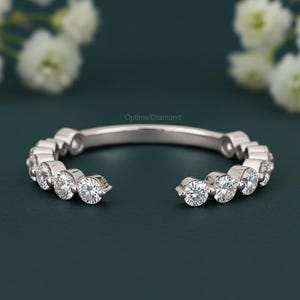 May include: A silver ring featuring a series of clear, round gemstones. The ring has an open design, with the gemstones set along the top curve. The background is a dark teal colour, with white flowers in the background.