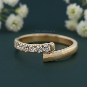 May include: A gold-colored ring with a unique open design. The ring features a row of small, sparkling diamonds set along one side. The ring is set against a dark teal background with white flowers.
