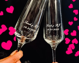 May include: Two clear glass champagne flutes with the text "Sarah & RJ 12.31.2023" etched on the front of each glass.