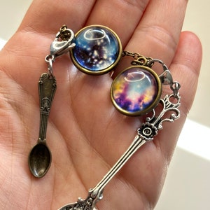 May include: A silver spoon with a decorative floral design on the handle and a round, scalloped bowl. The spoon is attached to a chain with two glass charms featuring galaxy-themed images. The charms are set in bronze-coloured metal.
