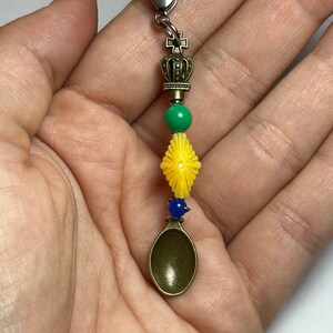 Puede incluir: Un charm decorativo con un pequeño colgante de cuchara de bronce. El charm incluye un cierre plateado, una corona, cuentas verdes y amarillas y una cuenta azul. El charm está diseñado para ser adjuntado a un llavero o cremallera.