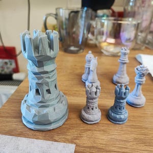May include: A collection of gray chess pieces, including a rook, knight, bishop, queen, and pawn, are arranged on a wooden surface. The rook is the largest piece, with a geometric design. The other pieces are smaller and have a similar color.