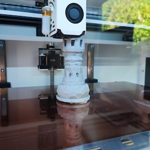 May include: A 3D printer in action, producing a white rook chess piece. The printer head is white with a black circular element. The rook is light grey and has a geometric design. The printer is set up in a well-lit environment.