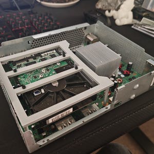 May include: An open, silver-colored electronic device with internal components visible. The device features a large fan, circuit boards, and a heat sink. The internal structure is visible, showcasing the intricate arrangement of the components.