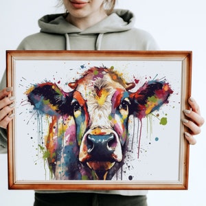 May include: A framed watercolor painting of a cow's head with colorful splashes of paint. The cow's face is mostly white with brown spots and the background is white.