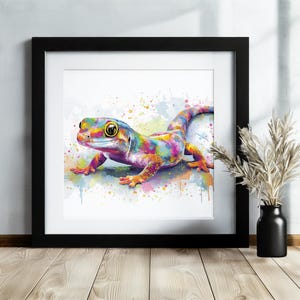 May include: A framed watercolor print of a gecko in vibrant colors. The gecko is depicted with a rainbow of hues, including yellow, blue, pink, and orange. The artwork is framed in a black square frame, adding a modern touch to the colorful reptile.