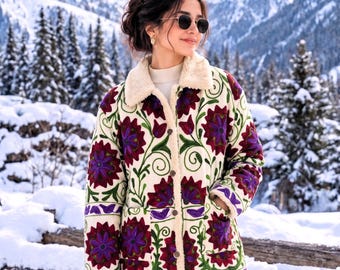 May include: A cream-colored jacket featuring a plush fleece lining and collar, adorned with vibrant purple and dark red floral embroidery and green vine patterns. The jacket includes a button-front closure and two front pockets, paired with cream-colored trousers and a small, fuzzy, cream-colored handbag with brown leather straps.