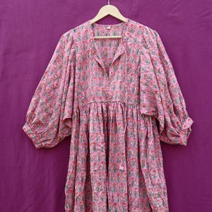 May include: A pink floral print dress with long, voluminous sleeves and a V-neckline. The dress features a gathered waist and a flowing skirt. The fabric is adorned with a repeating pattern of red and green floral motifs. The dress is hanging on a wooden hanger.