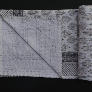 May include: A white and gray quilt with a leaf pattern and decorative border. The quilt is partially rolled up, revealing the intricate stitching and design. The overall aesthetic is simple and elegant.