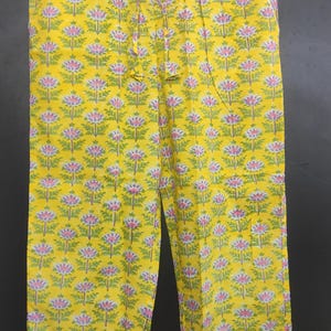 Op de afbeelding: Gele bloemenprint pyjamabroek. De broek heeft een herhalend patroon van roze en groene bloemen op een gele achtergrond. Het heeft een taille met trekkoord en witte bies langs de zakken en zoom. De broek hangt aan een houten hanger.