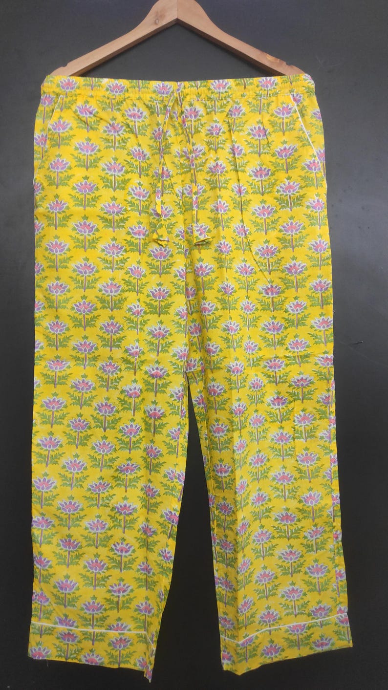 Op de afbeelding: Gele bloemenprint pyjamabroek. De broek heeft een herhalend patroon van roze en groene bloemen op een gele achtergrond. Het heeft een taille met trekkoord en witte bies langs de zakken en zoom. De broek hangt aan een houten hanger.