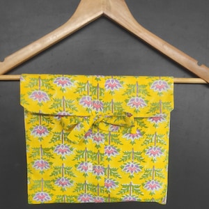 Op de afbeelding: Een opgevouwen geel stoffen item met een bloemenpatroon van paarse en groene bloemen, hangend aan een houten kleerhanger. De stof is vastgebonden met een bijpassend geel lint. De achtergrond is donkergrijs.