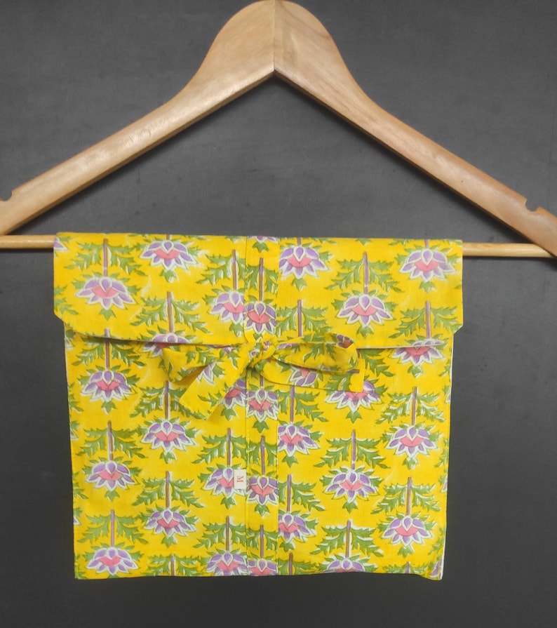 Op de afbeelding: Een opgevouwen geel stoffen item met een bloemenpatroon van paarse en groene bloemen, hangend aan een houten kleerhanger. De stof is vastgebonden met een bijpassend geel lint. De achtergrond is donkergrijs.