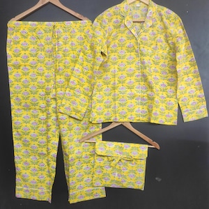 Op de afbeelding: Gele pyjamaset met een bloemenpatroon in paars en groen. De set bevat een overhemd met knopen, een broek en een bijpassend etui, allemaal hangend aan houten kleerhangers. De pyjama's hebben witte biezen.
