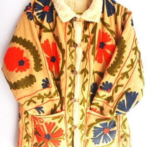 May include: A beige jacket with a faux fur collar and floral embroidery in shades of red, blue, and green. The jacket has two pockets and a button closure.