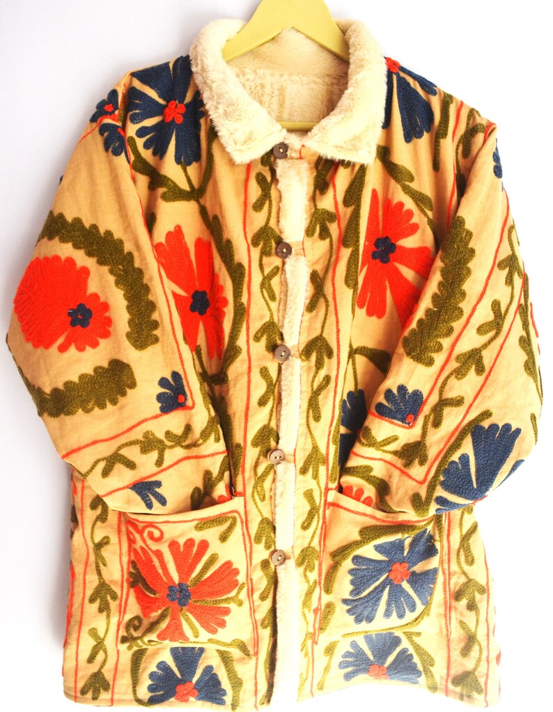 May include: A beige jacket with a faux fur collar and floral embroidery in shades of red, blue, and green. The jacket has two pockets and a button closure.