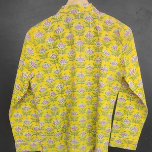 Op de afbeelding: Een gele shirt met lange mouwen met een bloemenpatroon. De shirt heeft een herhalend ontwerp van roze en paarse bloemen met groene bladeren. De shirt hangt aan een houten hanger.