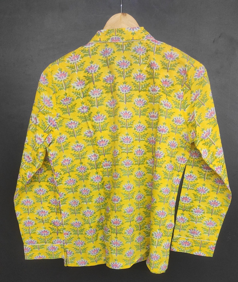 Op de afbeelding: Een gele shirt met lange mouwen met een bloemenpatroon. De shirt heeft een herhalend ontwerp van roze en paarse bloemen met groene bladeren. De shirt hangt aan een houten hanger.