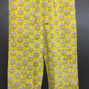 Op de afbeelding: Gele pyjamabroek met een bloemenpatroon in roze, groen en wit. De broek hangt aan een houten kleerhanger. Het bloemenpatroon herhaalt zich over de stof.